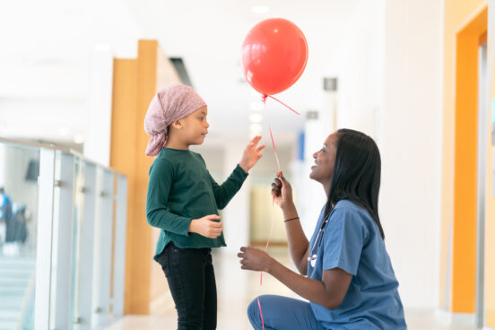 A beautiful African American girl with cancer is wearing a headscarf. She is in wearing the hospital corridor. Her kind female doctor is kneeling down and handing her a red ballon to cheer her up. The girl is reaching for the balloon with a smile.